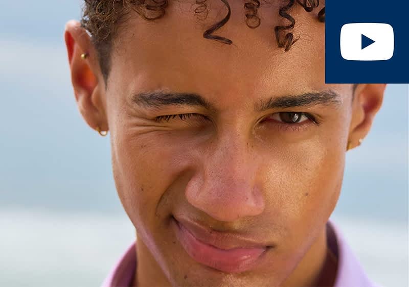junger Mann blickt in die Kamera, ein Auge zugekniffen, leichtes Stirnrunzeln, sichtbare Ohrpiercings, lockige Haare, unscharfer heller Hintergrund