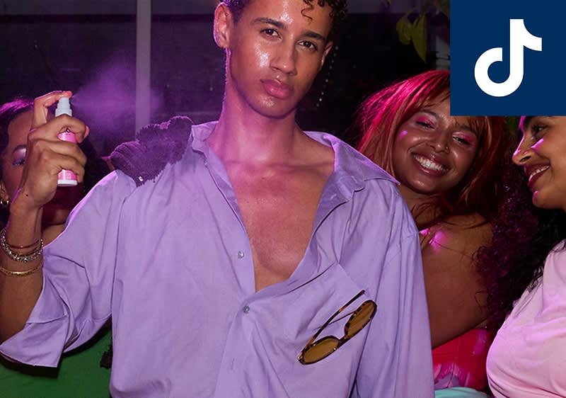 Mann mit offener hellvioletter Hemdbluse hält ein Spray in der Hand, Sonnenbrille steckt in der Brusttasche, im Hintergrund zwei weitere Personen bei Partybeleuchtung