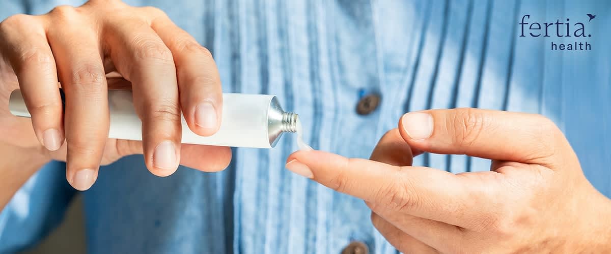 Hände drücken aus einer weißen Tube ein tröpfchenförmiges Gel auf einen Finger, im Hintergrund hellblaues Hemd und Markenlogo „fertia health“ sichtbar