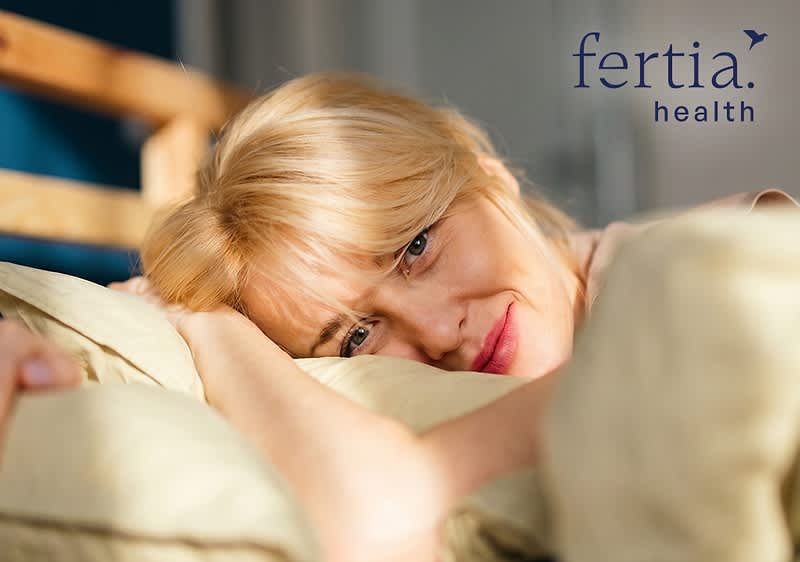 Frau liegt halb im Bett, stützt Kopf auf Kissen und blickt nach vorn, oben rechts blaues Logo und Schriftzug „fertia. health“