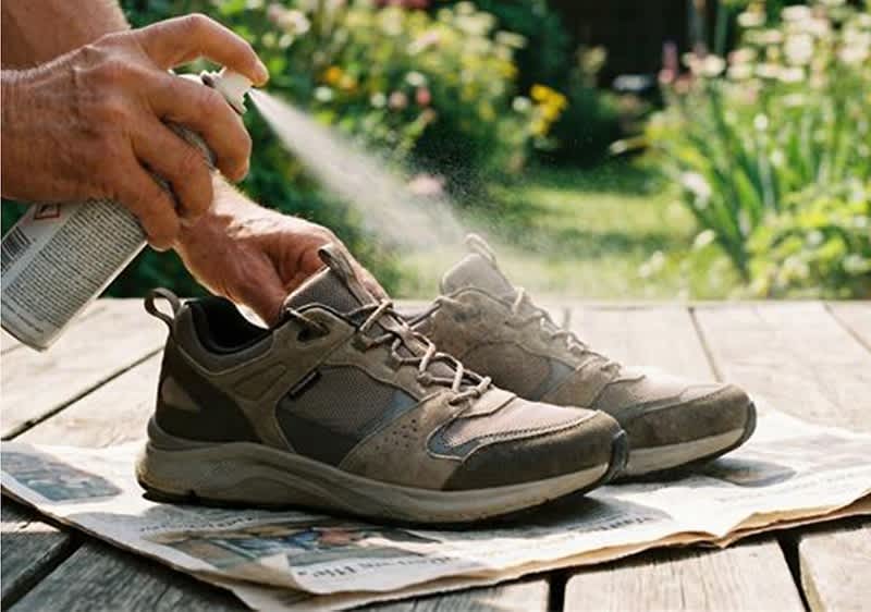 Hand sprüht Imprägnierspray auf ein Paar braune Outdoor-Sneaker, die auf ausgebreiteter Zeitung im Freien stehen