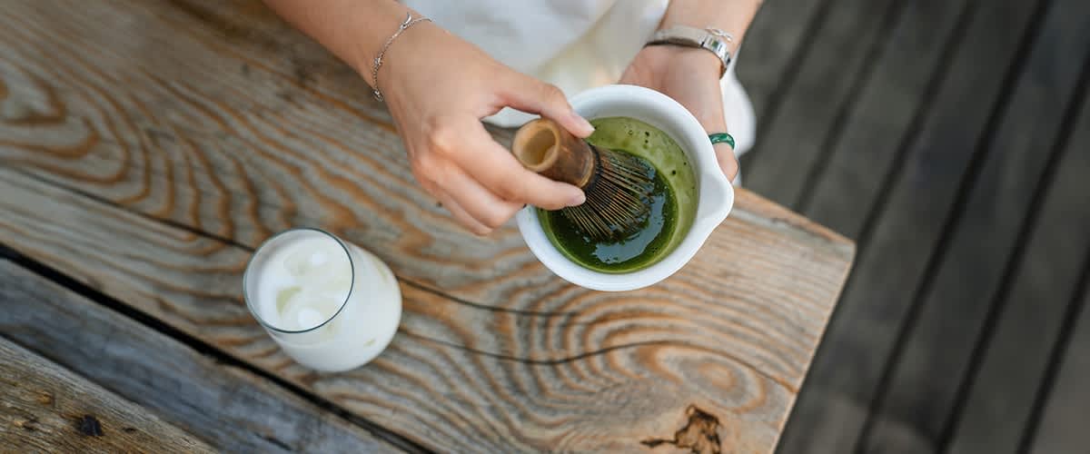 Person schäumt in einer weißen Schale grünes Matcha-Pulver mit einem Bambusbesen, daneben ein Glas mit Milch auf einem Holztisch