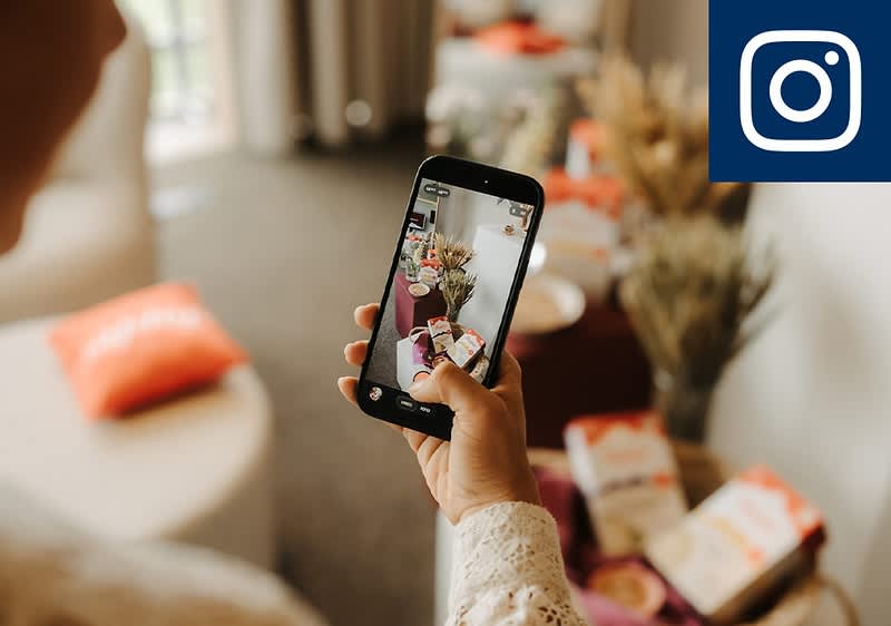 Hand hält Smartphone und fotografiert einen Tisch mit Snacks und Trockenblumen, oben rechts Instagram-Logo sichtbar