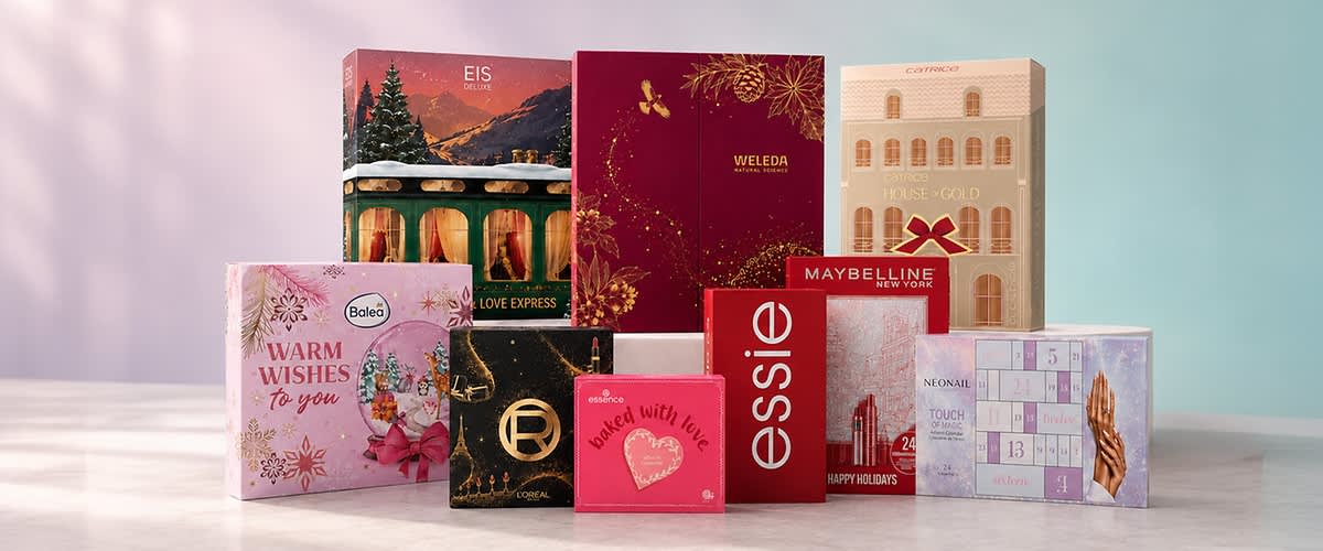 Verschiedene Adventskalender- und Geschenkboxen für Kosmetik und Pflege in unterschiedlichen Farben und Designs, darunter Boxen mit Markenlogos wie Essie, Maybelline und Weleda sowie verzierte Geschenkverpackungen mit Weihnachtsmotiven, auf hellem Untergrund ohne Personen