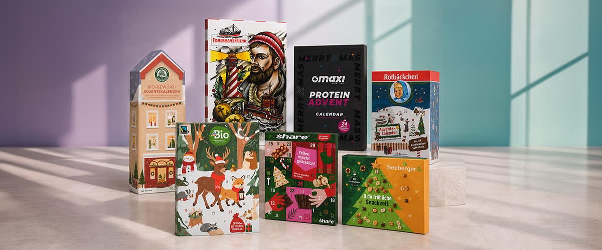 Verschiedene Adventskalender in Geschenkboxen, vorn links ein Bio-Gewürz-Adventskalender, daneben ein Kinder-Adventskalender mit Rentieren, mittig ein schwarzer „oMaxi Protein Advent Calendar“, rechts ein roten „Rotbäckchen“ Adventskalender und ein grüner Seeberger-Snack-Adventskalender sowie weitere bunte Adventskalender im Hintergrund. Bild mithilfe von KI erstellt