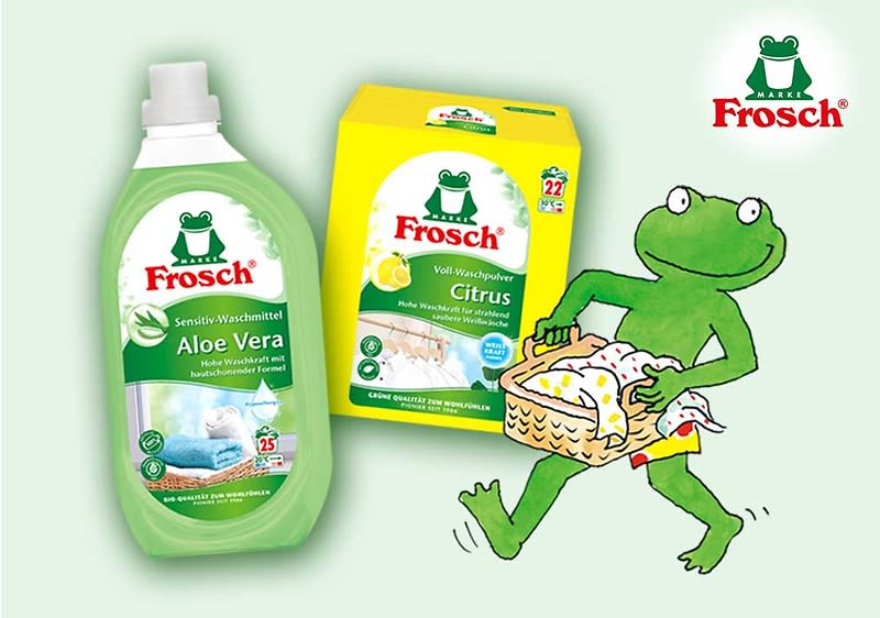 Zwei grüne Frosch-Marke Waschmittelverpackungen links: eine Flasche Frosch Sensitiv-Waschmittel Aloe Vera und eine gelbe Box Frosch Voll-Waschpulver Citrus, rechts eine grüne Cartoon-Froschfigur trägt einen Wäschekorb mit bunten Kleidungsstücken, oben rechts das Frosch-Logo