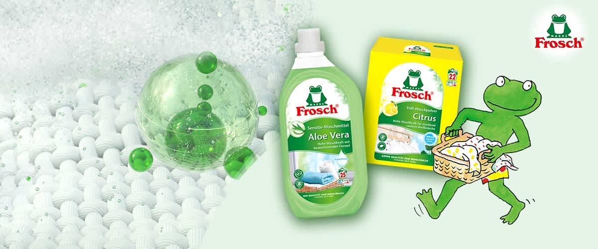 Zwei grüne Frosch-Marke Waschmittelverpackungen links: eine Flasche Frosch Sensitiv-Waschmittel Aloe Vera und eine gelbe Box Frosch Voll-Waschpulver Citrus, rechts eine grüne Cartoon-Froschfigur trägt einen Wäschekorb mit bunten Kleidungsstücken, oben rechts das Frosch-Logo