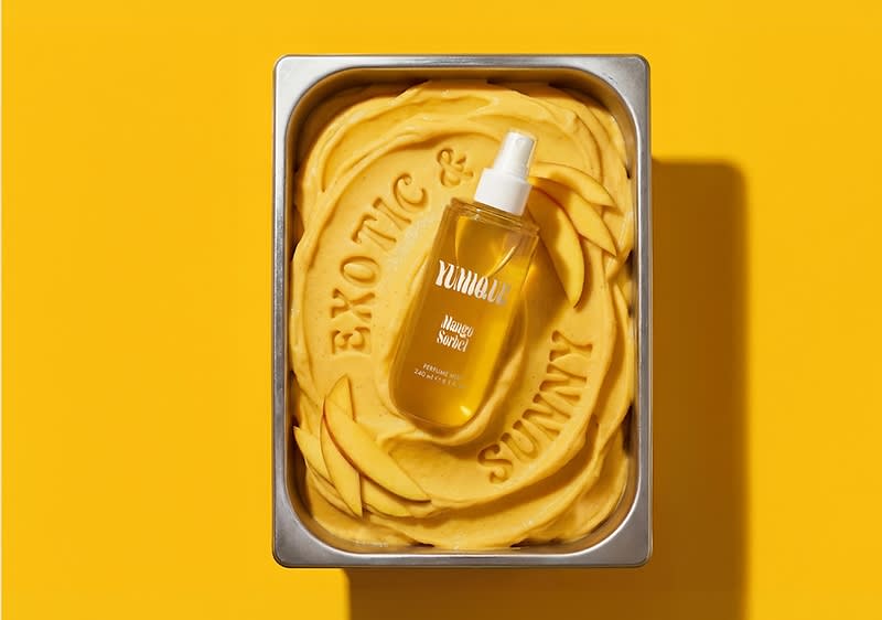 Glasflasche mit Pumpspray liegt in Metallwanne auf gelber Mango-Creme, eingeprägt die Wörter „EXOTIC & SUNNY“, Flaschenaufdruck „mango“ sichtbar