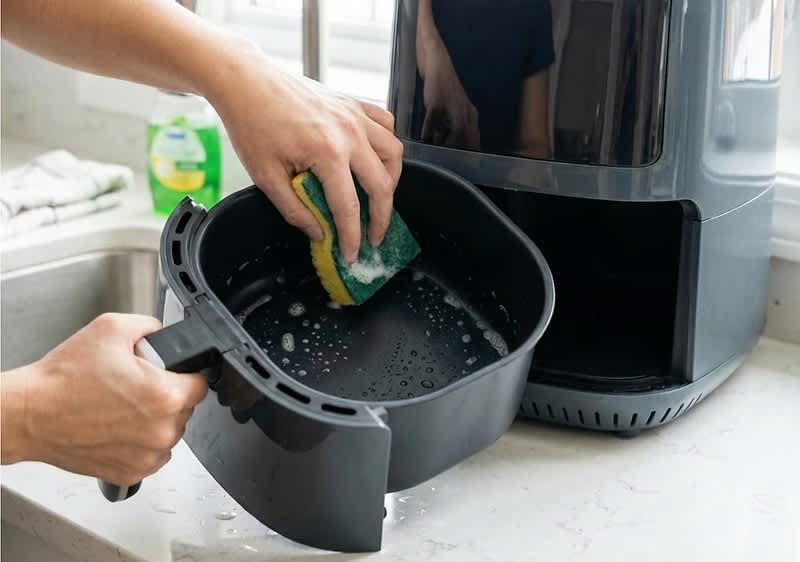Zwei Hände reinigen mit einem Schwamm den herausgenommenen Auffangbehälter einer Heißluftfritteuse, schaumiges Wasser im Behälter sichtbar