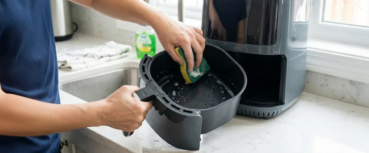 Zwei Hände reinigen mit einem Schwamm den herausgenommenen Auffangbehälter einer Heißluftfritteuse, schaumiges Wasser im Behälter sichtbar