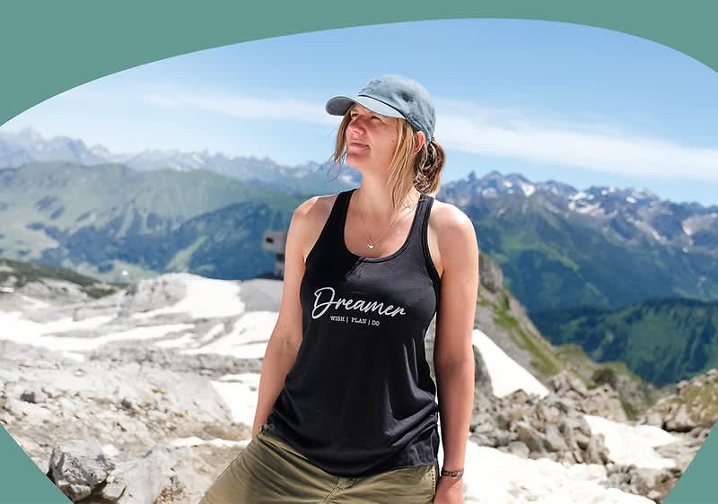 Frau in sportlichem Outfit mit schwarzem Tanktop mit weißem Schriftzug „Dreamer“ und khakifarbenen Shorts steht auf felsigem Berggipfel vor Alpenpanorama, trägt Kappe und leichtes Schmuckstück am Hals