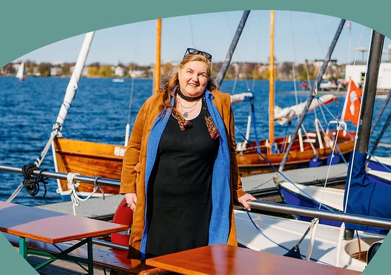 Petra Thomas steht auf einem Bootsdeck am Steg, trĂ€gt ein schwarzes oder Kleid mit offenem hellblauem Innenfutter und braunem Mantel darĂŒber, hĂ€lt sich an Reling fest; im Hintergrund Wasser und mehrere Segelboote sichtbar