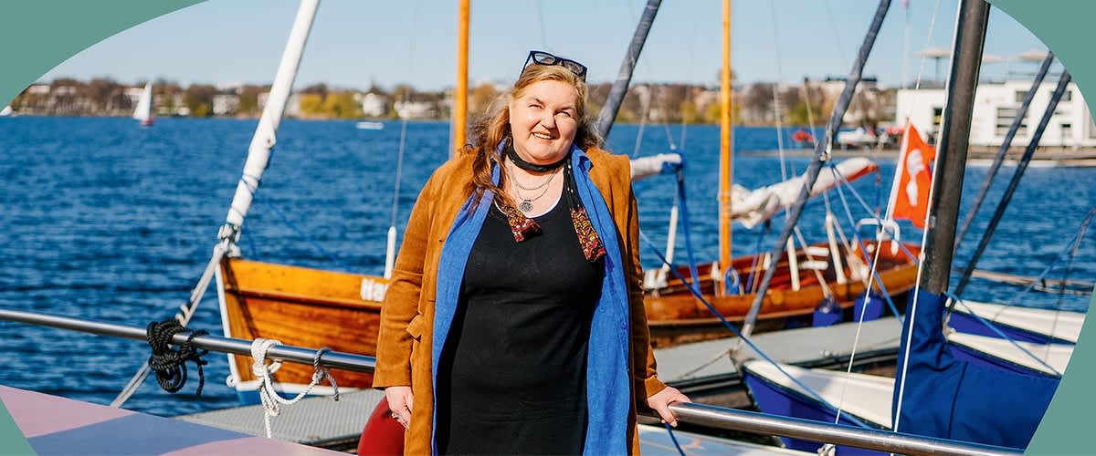 Petra Thomas steht auf einem Bootsdeck am Steg, trägt ein schwarzes oder Kleid mit offenem hellblauem Innenfutter und braunem Mantel darüber, hält sich an Reling fest; im Hintergrund Wasser und mehrere Segelboote sichtbar