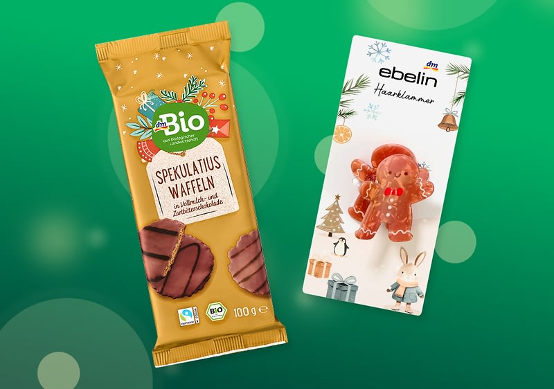 Zwei Produkte vor grünem Hintergrund: links eine goldgelbe Packung dmBio „Spekulatius Waffeln“ (100 g) mit Schokoladenüberzug, rechts eine ebelin Haarklammer in Form eines roten Lebkuchenmanns auf weißer Karte mit weihnachtlichen Illustrationen