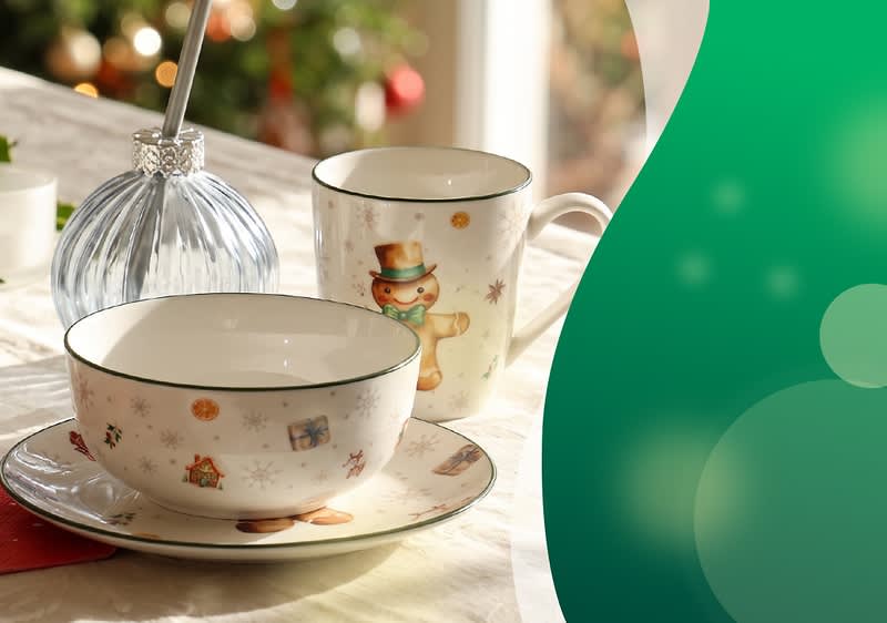 Porzellan-Tasse, Schale und Untertasse mit grüner Randlinie und weihnachtlichen Motiven (Schneemann, Lebkuchenhaus) auf Tisch, im Hintergrund Weihnachtsdeko. Bild mithilfe von KI erstellt