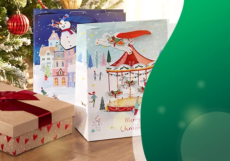 Zwei weihnachtliche Geschenktüten mit bunten Wintermotiven (Karussell und Schneemann) neben einem Geschenkpaket mit roter Schleife auf Holzboden. Bild mithilfe von KI erstellt