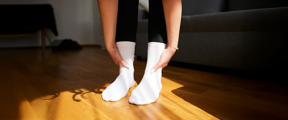 Person zieht weiße Sportsocken über beide Füße, Hände greifen die Socken am Schaft, Holzfußboden und Sofakante im Hintergrund