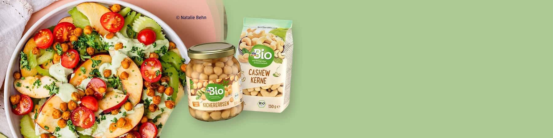 dmBio-Glas Kichererbsen und Packung Cashewkerne neben einem Salatteller mit Tomaten, Apfel- und Salatblättern auf grünem Hintergrund