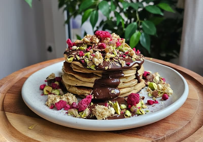 Stapel Pfannkuchen auf weißem Teller mit Schokoladensauce, Granola, gehackten Pistazien und getrockneten Himbeeren auf einem Holzuntersetzer