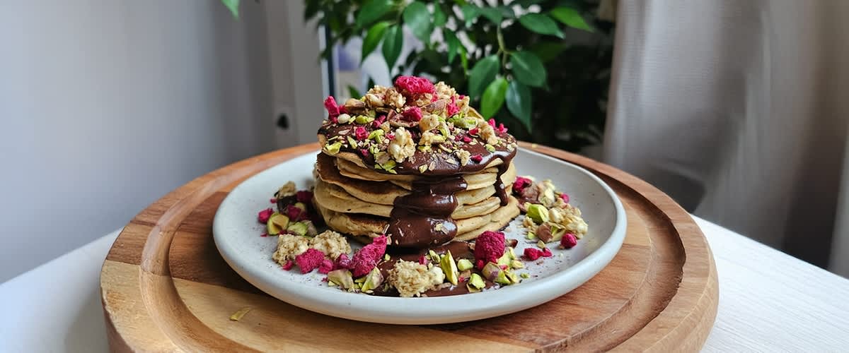 Stapel Pfannkuchen auf weißem Teller mit Schokoladensauce, Granola, gehackten Pistazien und getrockneten Himbeeren auf einem Holzuntersetzer
