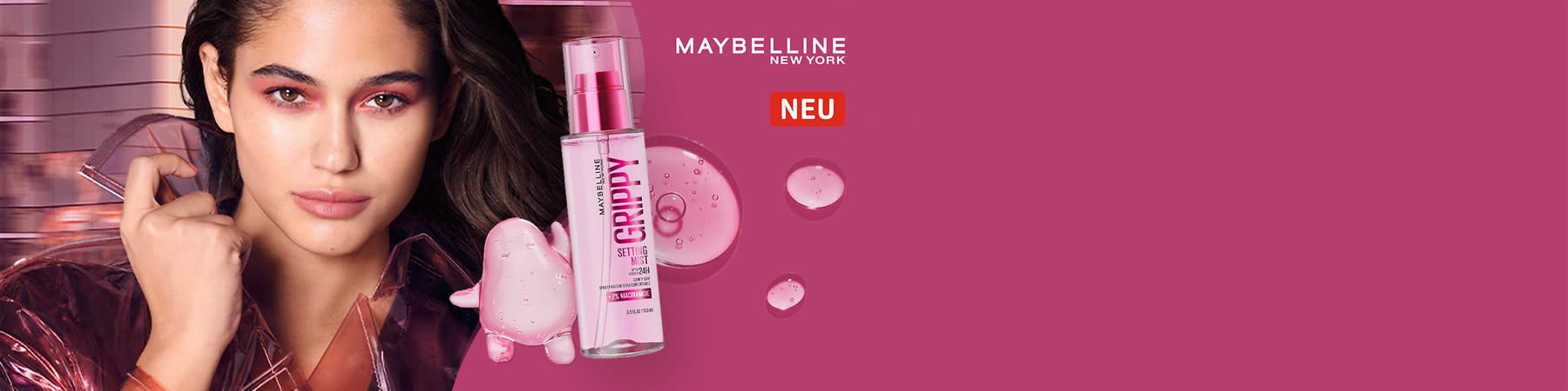 Maybelline Setting Mist „Grippy“ in durchsichtig-rosafarbener Sprühflasche, neben Produkttropfen auf pinkem Hintergrund und weißer Markenaufschrift „MAYBELLINE NEW YORK“ sowie rotem „NEU“-Label
