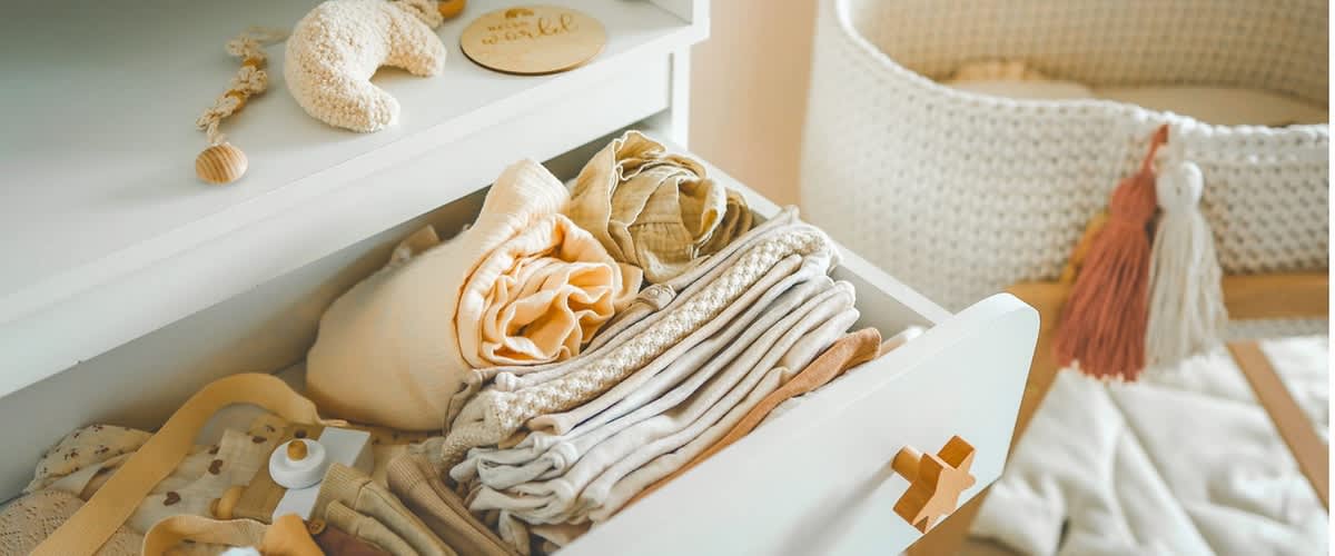 Offene Wickelkommode mit ordentlich gefalteter und gerollter Babykleidung in gedeckten Naturtönen, daneben ein geflochtener weißer Babykorb und eine Spielmatte mit Quasten