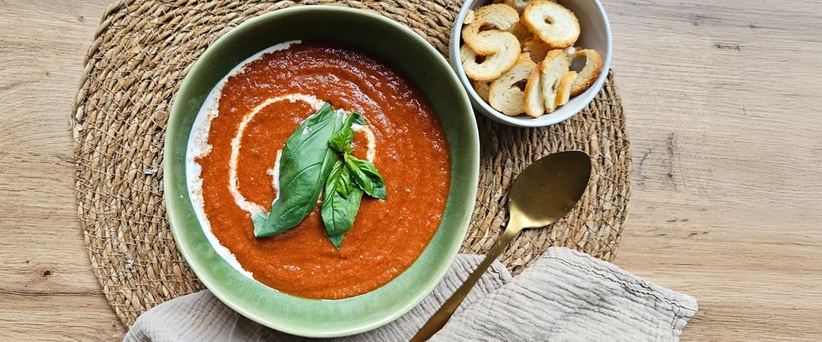 Grüne Schüssel mit roter Tomatensuppe, mit Sahneswirl und Basilikumblatt garniert, daneben Schälchen mit gerösteten Brotscheiben und goldener Löffel