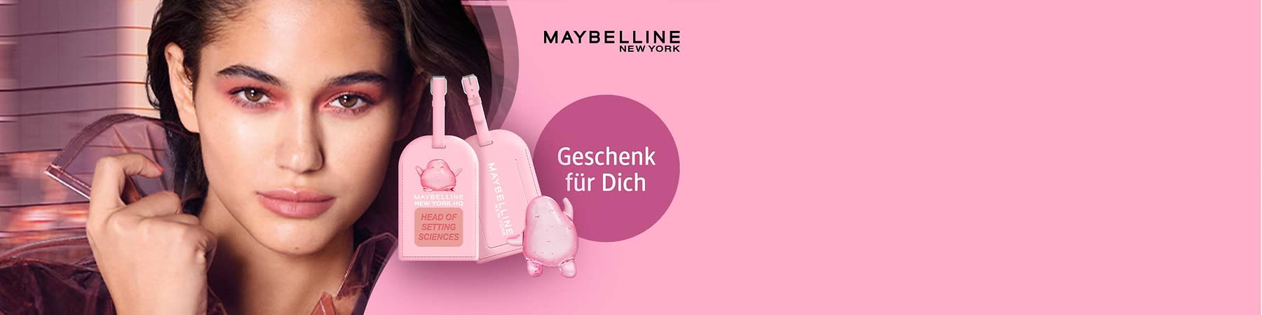 rosa Werbemotiv mit Maybelline New York Schriftzug oben rechts, zwei rosa Gepäckanhänger mit Aufschriften „MAYBELLINE NEW YORK HQ“ und „HEAD OF SETTING SCIENCES“ sowie eine runde violette Fläche mit weißer Aufschrift „Geschenk für Dich“, daneben eine kleine rosafarbene, glitzernde Figur ohne erkennbare Gesichtszüge