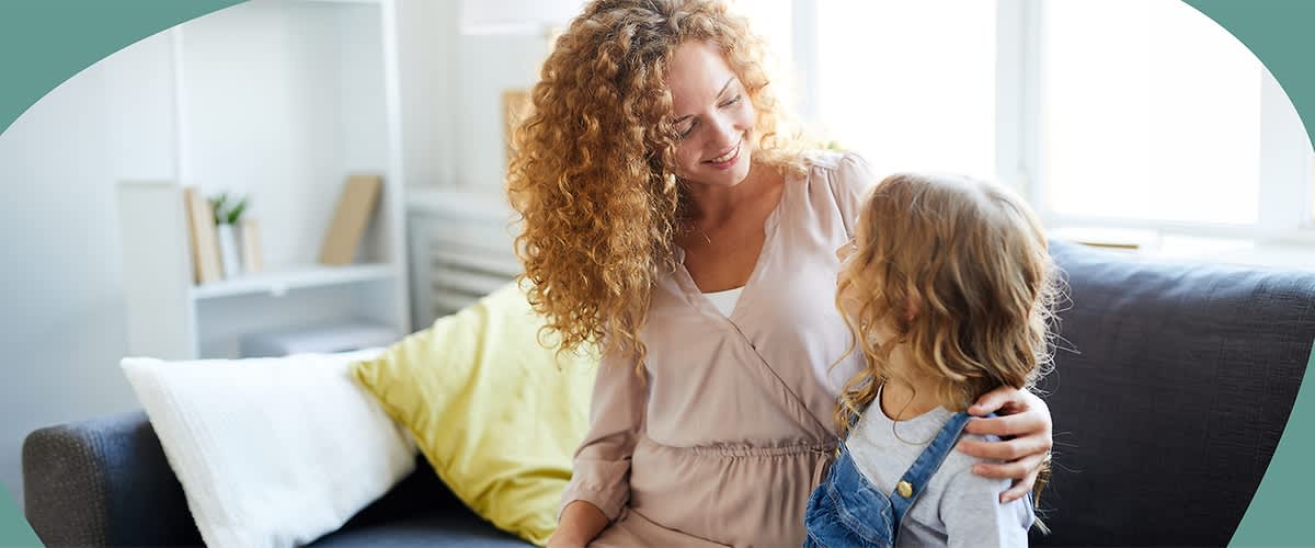 Zwei sitzende Personen auf einem Sofa: eine Erwachsenenperson mit lockigen roten Haaren legt den Arm um ein Kind mit schulterlangen blonden Locken und beide lächeln sich an