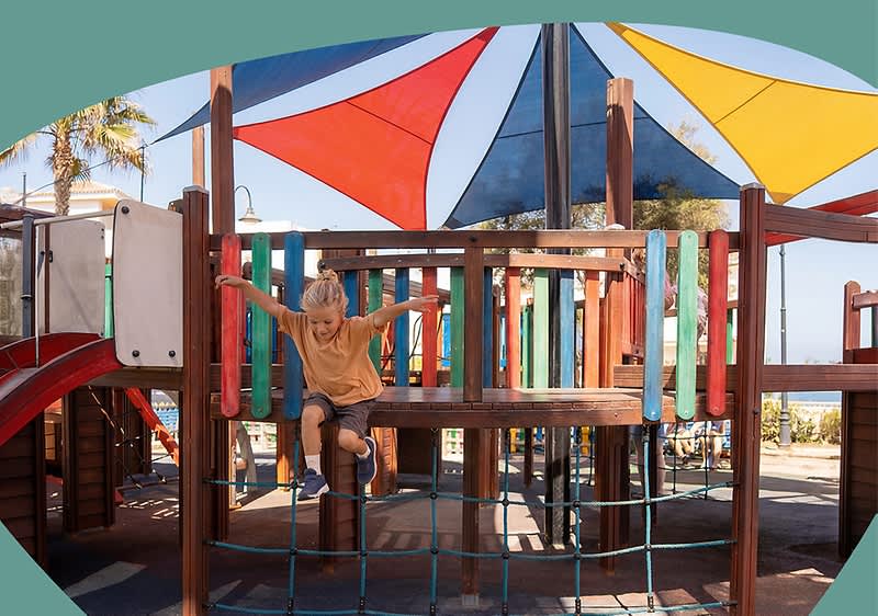 Kindes auf einem Holzklettergerüst eines Spielplatzes, das von bunten Sonnensegeln überdacht ist, Kind hält sich an Geländern fest und steht auf einem Netzbalancierweg. Bild mithilfe von KI erstellt