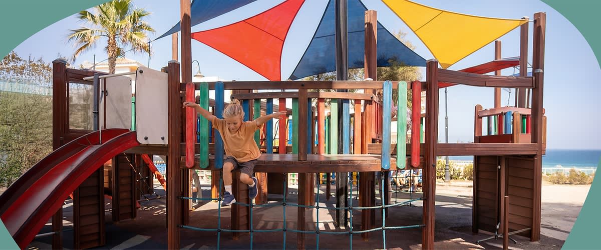 Kindes auf einem Holzklettergerüst eines Spielplatzes, das von bunten Sonnensegeln überdacht ist, Kind hält sich an Geländern fest und steht auf einem Netzbalancierweg. Bild mithilfe von KI erstellt