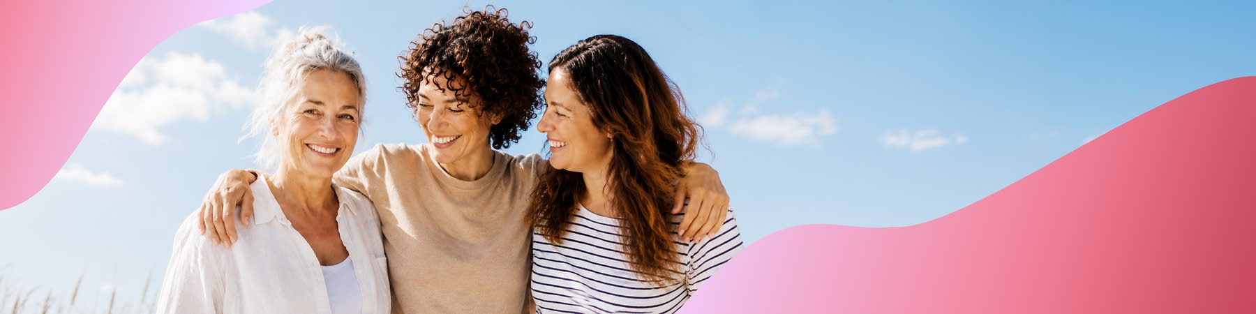 Drei Frauen verschiedener Altersstufen stehen eng beieinander, blicken zueinander und lächeln, die mittlere Frau hat den Arm um die beiden anderen gelegt, blauer Himmel im Hintergrund