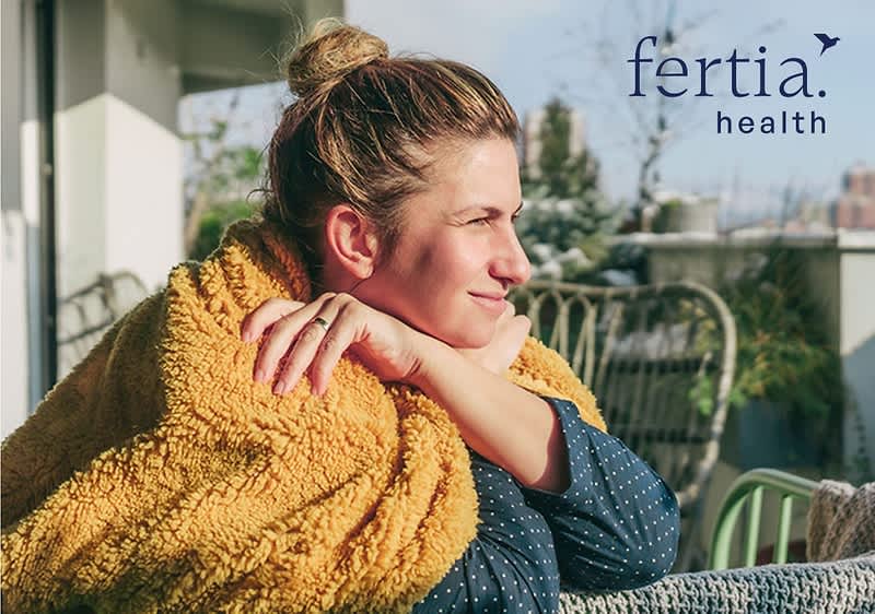 Frau sitzt auf Balkon, in Decke gehüllt, stützt Kopf auf verschränkte Arme und blickt nach rechts; Markenlogo „fertia health“ oben rechts