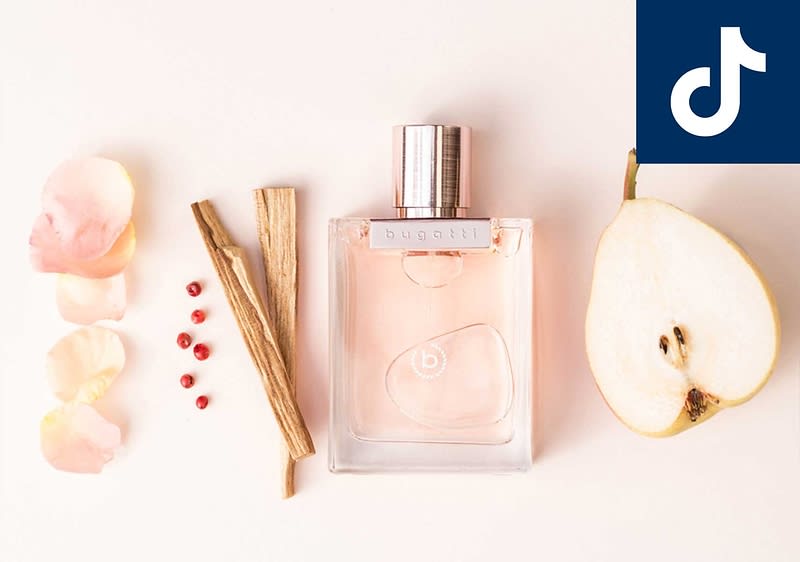 Parfümflakon der Marke bugatti mit rosafarbenem Inhalt, links Rosenblütenblätter, rosa Pfefferkörner und zwei Holzstäbchen, rechts halbierte Birne und TikTok-Logo oben rechts