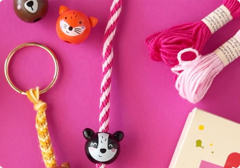 rosa Hintergrund mit verschiedenen Bastelmaterialien: geflochtenes Schlüsselband mit goldenem Schlüsselring, pink-weiß gedrehtes Band mit schwarzem Bärenperlenanhänger, zwei pinke Garnknäuel und zwei runde Holzperlen mit Tiergesichtern, rechts unten Ecke einer bunten Verpackung