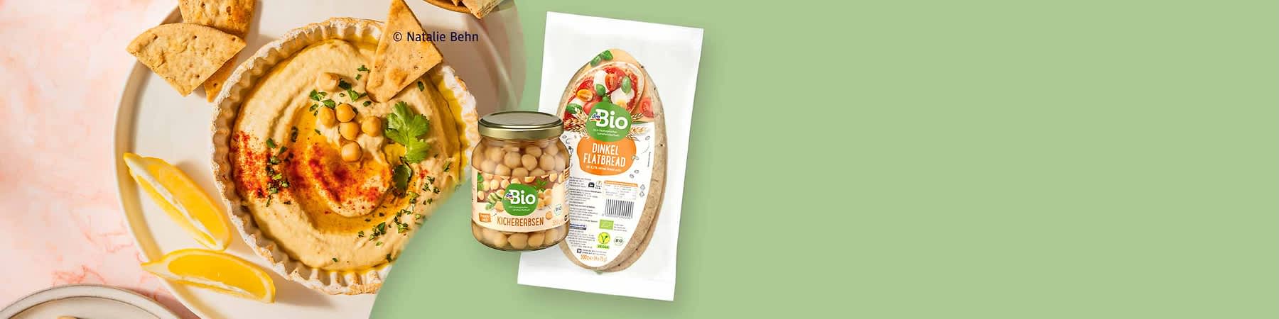 Glas mit Bio-Kichererbsen neben einer weiß-grünen Verpackung mit Aufschrift „Bio Dinkel Flatbread“, im Hintergrund Hummus mit Kichererbsen, Kräutern, Tortillastücken und Zitronenspalten