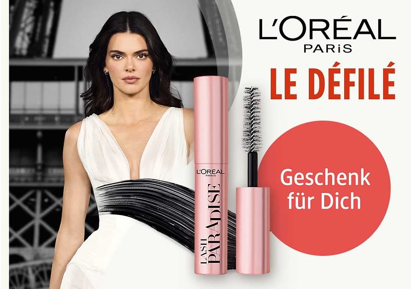 Roségoldene L'Oréal Lash Paradise Mascara mit geöffnetem Bürstenkopf, schwarze Mascara-Striche links, roter Kreis mit Text „Geschenk für Dich“ und Schriftzug „L'ORÉAL PARIS LE DÉFILÉ“ oben rechts