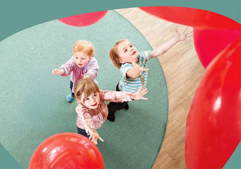 Drei Kinder auf einem grünen Teppich greifen nach mehreren roten Luftballons, Blick von oben auf die Szene