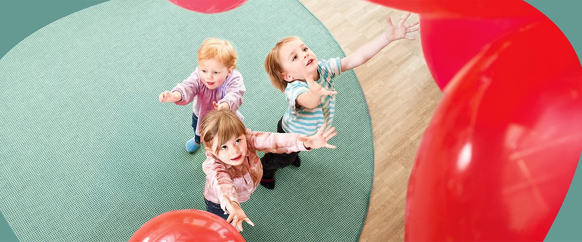 Drei Kinder auf einem grünen Teppich greifen nach mehreren roten Luftballons, Blick von oben auf die Szene