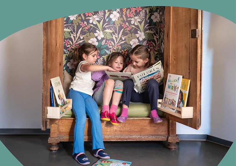 Drei Kinder sitzen in einem offenen Schrank und lesen gemeinsam Bilderbücher, zwei Bücherständer mit weiteren Kinderbüchern stehen an den Schranktüren