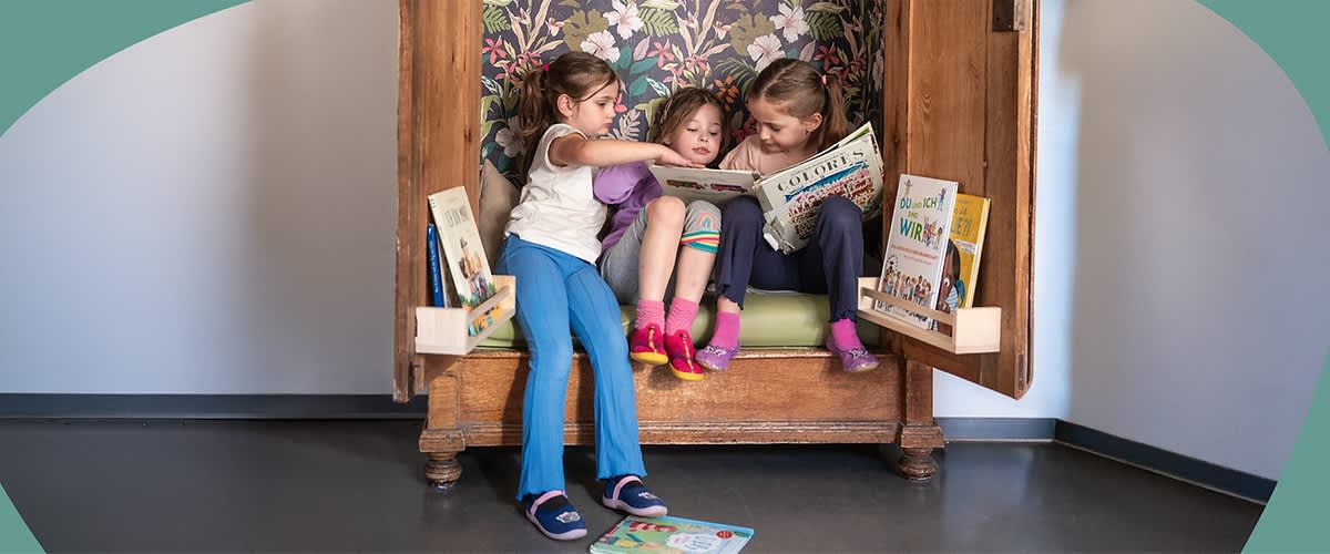 Drei Kinder sitzen in einem offenen Schrank und lesen gemeinsam Bilderbücher, zwei Bücherständer mit weiteren Kinderbüchern stehen an den Schranktüren