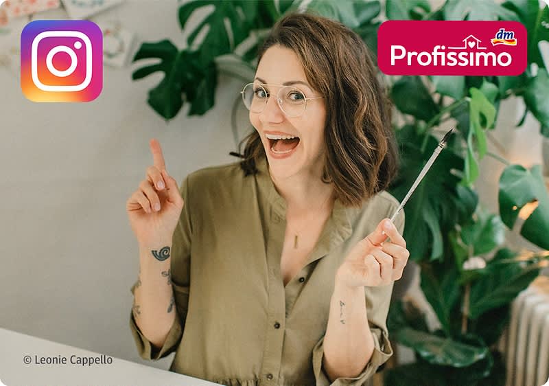 junge Frau mit Brille lächelt und zeigt mit einem Finger nach oben, in der anderen Hand hält sie einen langen Make-up-Pinsel, Hintergrund mit großen grünen Zimmerpflanzen, oben rechts Logo „Profissimo“, oben links Instagram-Logo