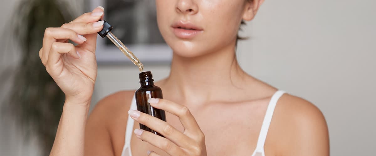 Frau hält braune Glasflasche und Pipette, mit Tropfen eines Öls über der Flaschenöffnung