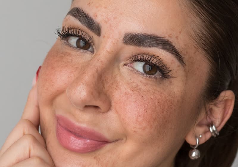 Nahaufnahme eines lächelnden Gesichts mit Sommersprossen, betonten dunklen Augenbrauen, langen Wimpern, rosa Lippen und mehreren silbernen Ohrringen am rechten Ohr