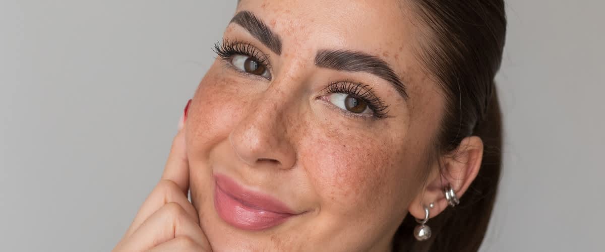 Nahaufnahme eines lächelnden Gesichts mit Sommersprossen, betonten dunklen Augenbrauen, langen Wimpern, rosa Lippen und mehreren silbernen Ohrringen am rechten Ohr