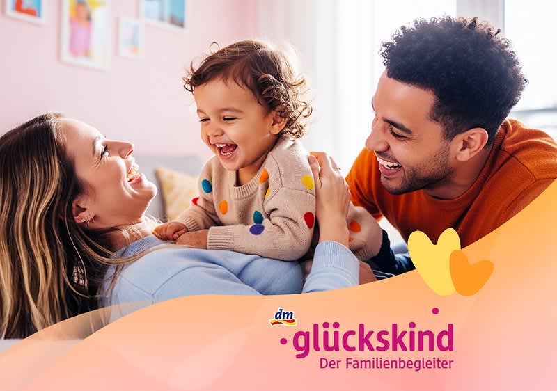 Eine lächelnde Elternpaar liegt gegenüber und hält ein lachendes Kleinkind auf dem Bauch, darunter Schriftzug glückskind Der Familienbegleiter und dm-Logo auf rosa-orangenem Wellenhintergrund. Bild mithilfe von KI erstellt