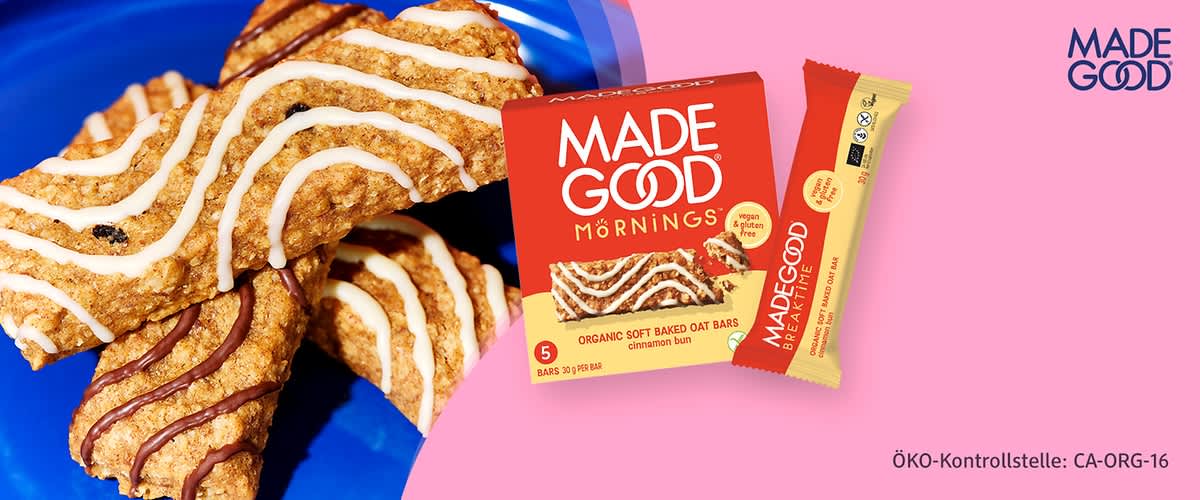Rote Verpackung der Marke MADE GOOD Mornings mit Bild eines Zimt-Hafer-Riegels; daneben ein einzelner beige-roter Riegel mit der Aufschrift „MADEGOOD“ und Hinweis „organic soft baked oat bars cinnamon bun“ sowie Text „ÖKO-Kontrollstelle: CA-ORG-16“