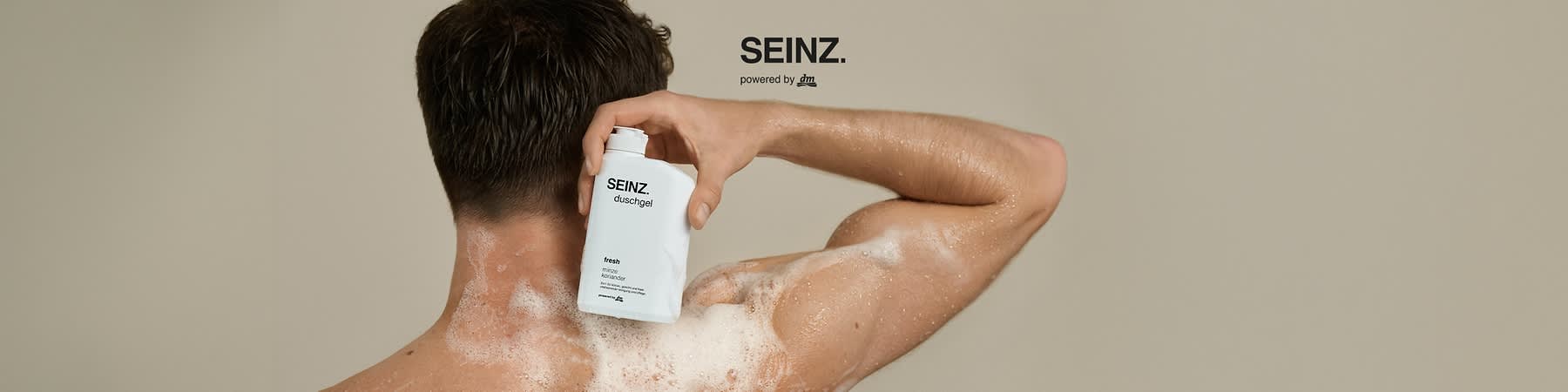 Mann von hinten unter der Dusche, schaumiger Rücken, Hand hält eine weiße Flaschenpackung mit Aufschrift „SEINZ. duschgel fresh minze koriander“ und aufgedrucktem dm-Logo, großer Schriftzug „SEINZ.“ oben rechts