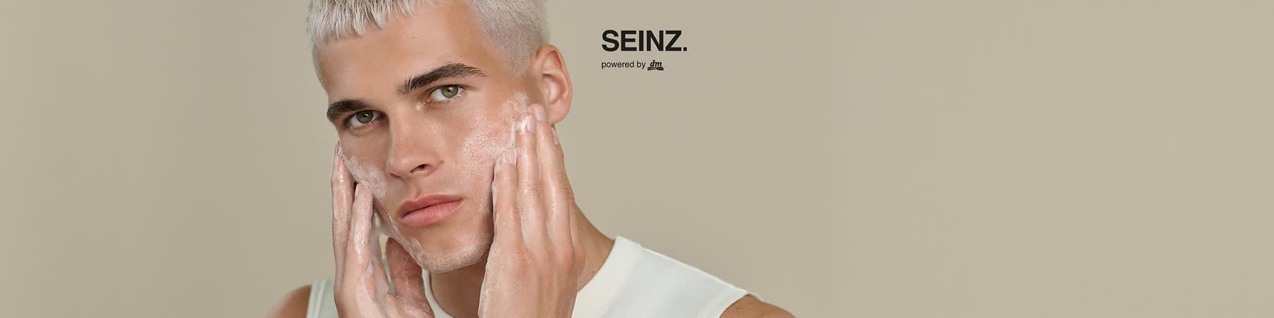Mann mit kurz platinblondem Haar trägt Gesichtsreinigungsschaum auf, beide Hände am Gesicht, rechts oben Schriftzug „SEINZ. powered by dm“