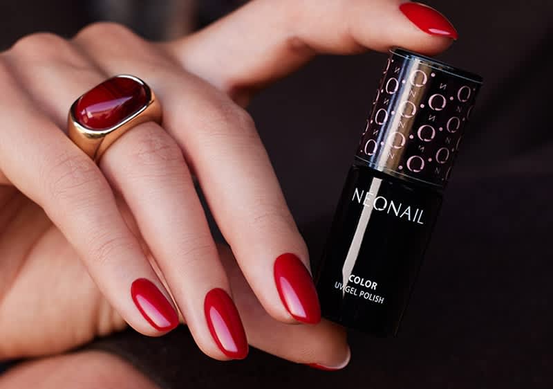 Hand mit ovalen, rot lackierten Fingernägeln hält eine schwarze Nagellack-Flasche mit der Aufschrift „NEONAIL Color UV Gel Polish“ und einen goldenen Ring mit rotem Stein